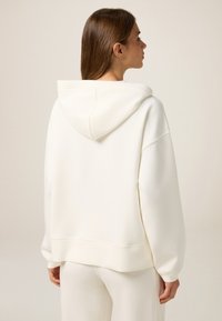 Sudadera crema con una gran capucha, corte relajado y puños acanalados. Tejido suave y diseño minimalista, sin patrones ni logotipos visibles.