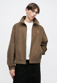 Brun kordfløyelsjakke med ståkrage, glidelås, ribbestrikkede mansjetter og subtil brodering på brystet, parret med en hvit skjorte og svarte bukser.