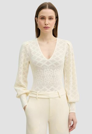 Donna che indossa un top a maglia color crema con scollo a V, maniche lunghe arricciate e pantaloni color crema a vita alta su uno sfondo grigio semplice.