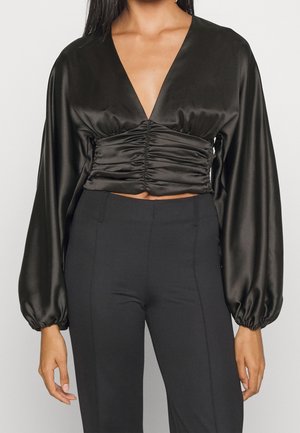 Vrouw draagt een zwarte satijnen cropped top met lange mouwen en plooien, met een diepe V-hals, gecombineerd met hoog getailleerde zwarte broek.
