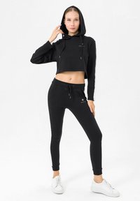 Felpa nera cropped con cappuccio, lacci e logo, abbinata a pantaloni neri aderenti con strisce laterali e tasche, indossata con sneakers bianche.