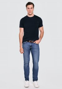 Mężczyzna ubrany w czarny t-shirt z krótkim rękawem, niebieskie dżinsy z brązowym paskiem i białe trampki, stojący na jednolitym tle.