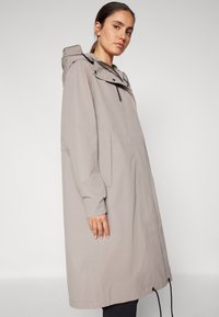 Cappotto impermeabile grigio chiaro con cappuccio, maniche lunghe, tasche frontali e orlo con cordoncino; texture liscia e design minimalista.