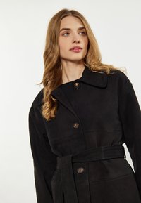 Giacca in suede nera con un ampio colletto, chiusura con bottoni e cintura in tessuto coordinato; texture liscia con sottili dettagli di cucitura.
