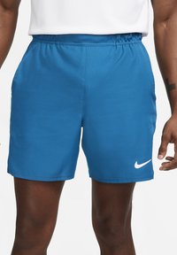 Shorts de sport bleus avec taille élastique et poches latérales portés par une personne à la peau foncée avec un tatouage sur l'avant-bras.