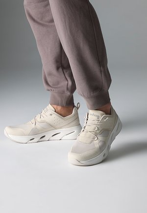 Person wearing beige athletic sneakers and mauve jogger pants, standing on a smooth light gray surface.