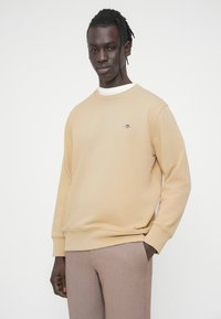 Sweat-shirt beige avec encolure et poignets côtelés, arborant un petit logo brodé sur la poitrine. Associé à un pantalon marron clair sur mesure.
