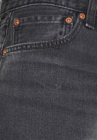 Gros plan sur la poche d'un jean en denim noir avec des rivets en cuivre visibles et des coutures détaillées.