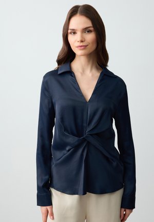 Blusa a maniche lunghe blu navy con colletto, dettaglio frontale intrecciato e tessuto liscio e lucido. Abbinata a pantaloni beige chiaro.