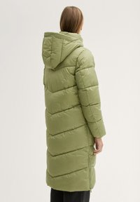 Parka verde largo y hinchado con capucha. Presenta secciones acolchadas horizontales y bolsillos con cremallera. Textura suave con un ligero brillo.
