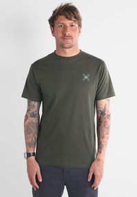 Camiseta de algodón verde oliva con mangas cortas, cuello redondo, que presenta un pequeño gráfico blanco en el área del pecho izquierdo. Suave, de ajuste estándar.