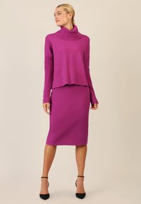 Suéter púrpura con un alto cuello tipo cowl y detalles de manga acanalada, combinado con una falda lápiz de longitud hasta la rodilla en el mismo tono y zapatos de tacón negros.