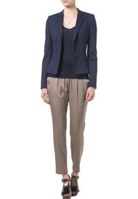 Kobieta ubrana w granatową marynarkę, czarny top, beżowe spodnie i czarne sandały na obcasie z frędzlami, stojąca na białym tle.