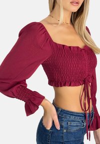Top court bordeaux à manches bouffantes, corsage froncé, ourlet volanté et fermeture à nouer. Porté avec un jean bleu taille haute.