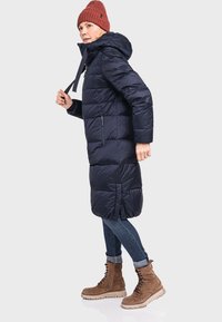 Marineblaue Steppjacke mit Kapuze, ausgestattet mit einem Reißverschluss vorne und Kordelzug, kombiniert mit blauen Jeans und braunen Schnürstiefeln bis zur Knöchel.