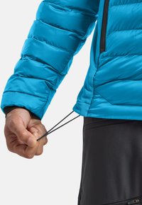 Giacca blu gonfiabile in materiale sintetico, dotata di tasca con zip e cordoncino regolabile all'orlo, indossata sopra pantaloni neri.