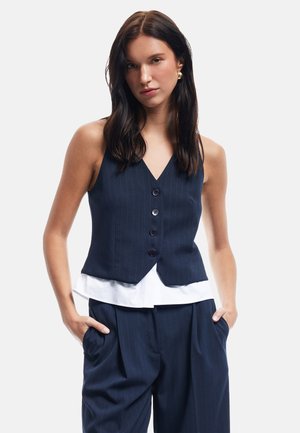 Gilet bleu marine à rayures avec cinq boutons, design en V, superposé sur une chemise blanche. Fabriqué en tissu lisse avec une coupe cintrée.