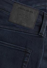 Jean en denim bleu foncé avec une étiquette en cuir embossée de texte, incluant "JACK & JONES", et une surface lisse et texturée.