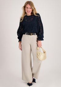 Marineblauwe kanten blouse met ballonmouwen, beige wijde broek, gouden riem en klein gouden geweven handtasje. Dikke marineblauwe strik als accent op de schoenen.