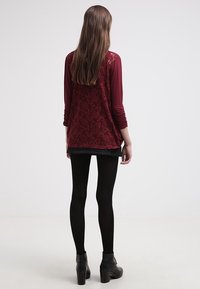 Burgundská halenka s krajkovými detaily na rukávech, černá mini sukně a černé punčocháče. Modelka na sobě má robustní boty na podpatku, pohled zezadu.