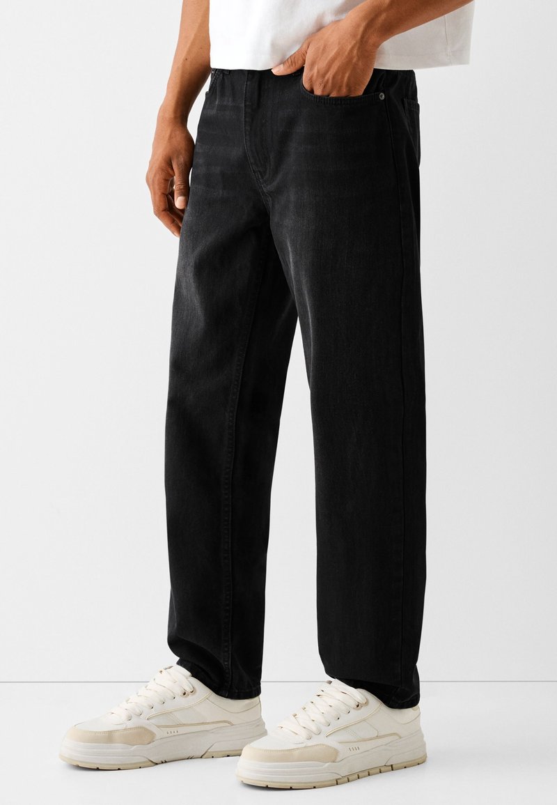 Jeans de mezclilla negra con corte recto, costuras detalladas y bolsillos delanteros, combinados con zapatillas crema. Textura suave, diseño casual.