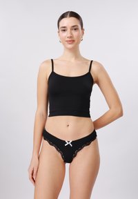Γυναίκα στέκεται φορώντας μαύρο κοντό τοπ και μαύρο εσώρουχο με δαντέλα και μικρό λευκό φιόγκο μπροστά.