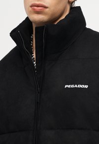 Nahaufnahme einer Person, die eine schwarze Zip-up-Jacke mit hohem Kragen und weißem "PEGADOR"-Logo auf der Brust trägt, wobei das untere Gesicht und ein Ohrring zu sehen sind.