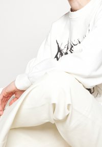 Vit sweatshirt med svart grafisk design, i kombination med krämfärgade byxor. Texturerad tyg synlig, avslappnad passform, visad i sittande position.