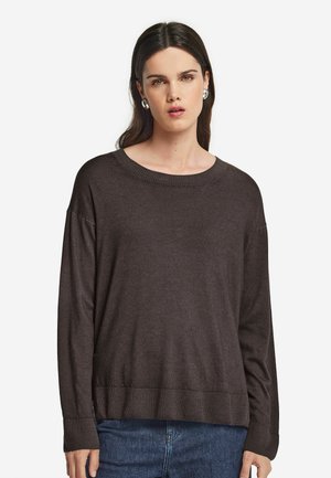Kvinde med langt mørkt hår iført sølvfarvede creoler, en mørkebrun trøje med lange ærmer og blå jeans, vendt mod fronten mod en hvid baggrund.
