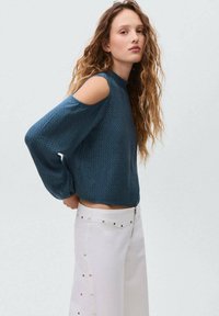 Bluză lungă de jerse cu mâneci lungi, de culoare teal, cu umeri decupați, model texturat, guler înalt și croială lejeră, asortată cu pantaloni albi cu talie înaltă, decorati cu piese metalice.