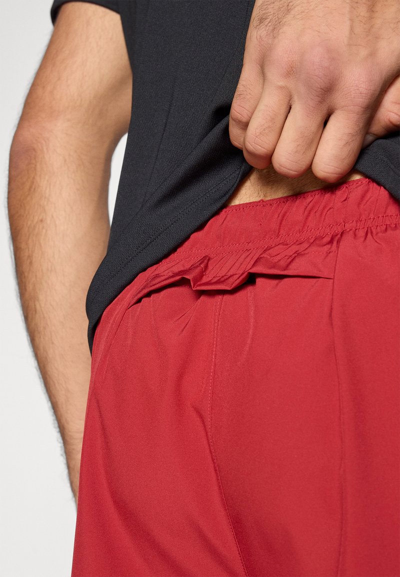 Pantaloncini atletici rossi con una cintura elastica, tessuto strutturato e un dettaglio arricciato nella parte superiore. Abbinati a una maglietta scura in vita.