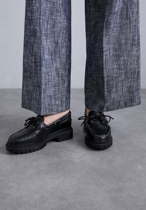 Black leather boat shoes with a thick rubber sole, detailed stitching, and lace-up design, paired with wide, textured gray trousers.