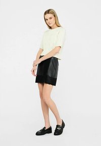 Maglione a maniche corte color crema con lavorazione a trecce abbinato a una gonna in ecopelle nera con orlo in pizzo smerlato, indossato con mocassini neri.