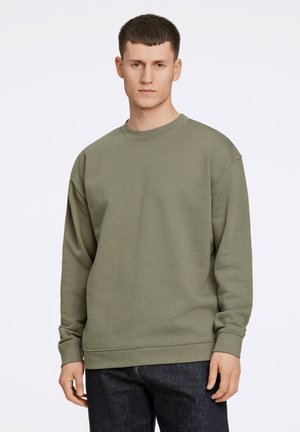 Felpa in cotone verde oliva con maniche lunghe, collo a girocollo ribattuto e orli a coste, indossata sopra jeans blu scuro. Il tessuto appare morbido e strutturato.