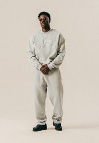 Mężczyzna w szarym bluzie BLKVIS, pasujących dresach, czarnych butach i okularach, stojący na jednolitym jasnym tle.