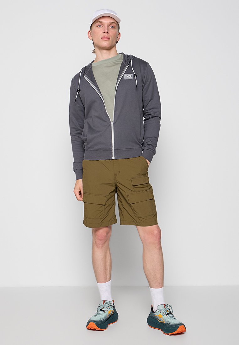 Felpa grigia con cappuccio e zip, t-shirt verde oliva e pantaloni cargo. Il modello indossa sneakers blu-grigie con accenti arancioni e calze bianche.