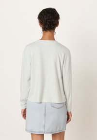 Kvinna med lockigt hår i en knut, som bär små hoop-örhängen, en ljusgrå långärmad topp och en ljus jeanskjol, stående mot en enfärgad bakgrund.