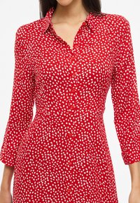 Femme portant une robe rouge à pois blancs irréguliers, fermeture à boutons sur le devant et manches trois-quarts, vue de l'épaule à la hanche.