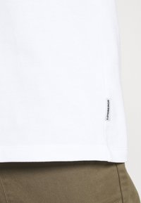Camiseta blanca de algodón con dobladillo recto, que presenta una pequeña etiqueta lateral con "LINDENBERGH" en texto negro. Textura lisa, sin patrones.