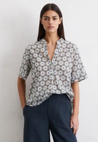 Blouse à manches courtes avec col, ornée d'un motif floral blanc et marron sur un fond clair. Fabriquée en tissu léger.