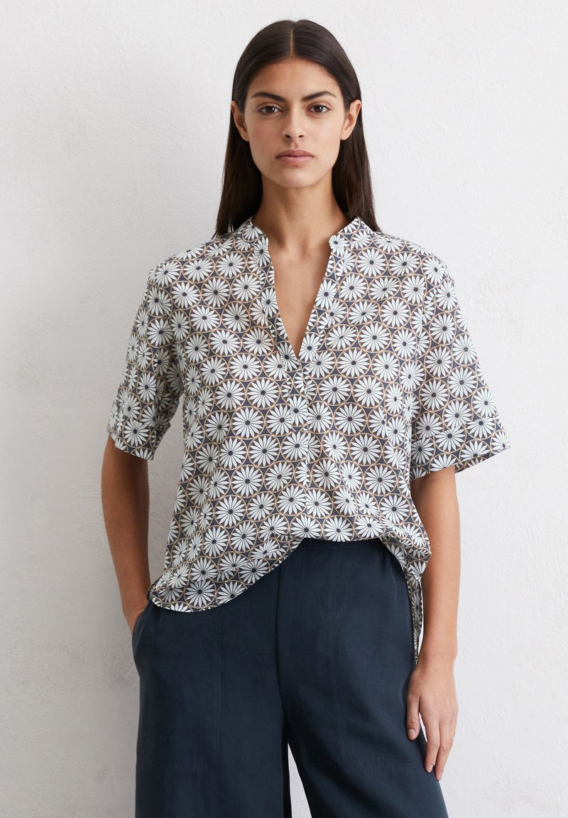 Blouse à manches courtes avec col, ornée d'un motif floral blanc et marron sur un fond clair. Fabriquée en tissu léger.