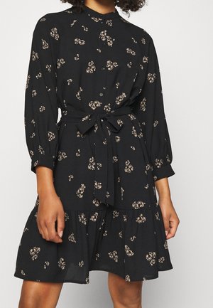 Vrouw draagt een zwarte, lange mouw, jurk met knopen, met kleine beige bloemmotieven en een ceintuur met strik in de taille.