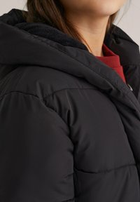 Sieviete, tērpta melnā izolētā ziemas jakā ar lielu kapuci, zem kuras redzama sarkana adīta džemperis; attēlota no pleca līdz zodam.