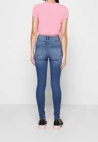 Mujer vestida con una camiseta corta de manga corta rosa, jeans ajustados de cintura alta azul y zapatos de plataforma negros con suelas rosas, de pie de espaldas.