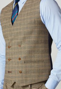 Muž v hnedo-károvanom dvojradovom vestu cez modrú košeľu s pruhovanou námořníckou modrou a červenou kravatou, s rukami v vreckách.