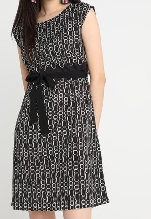 Vestito nero senza maniche con motivo a catena bianca, fiocco nero in vita, indossato da persona con capelli lunghi e scuri e orecchini colorati con nappine.