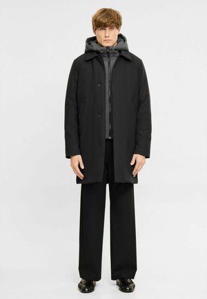 Joven de pie, vestido con un abrigo negro sobre una chaqueta gris con capucha, pantalones negros de pierna ancha y zapatos negros sobre un fondo liso.