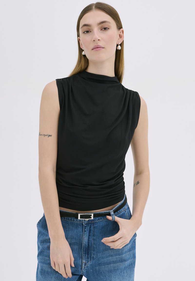 Haut sans manches noir avec un col montant, présentant un tissu drapé et une texture douce. Associé à un jean en denim bleu clair et une ceinture simple.