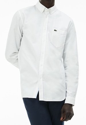 Homme portant une chemise blanche à boutons avec un petit logo de crocodile vert sur la poche poitrine, associée à un pantalon foncé.