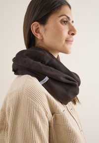 Femme portant une veste en velours côtelé beige et une écharpe tour de cou marron foncé, tournée vers la droite sur un fond clair uni.